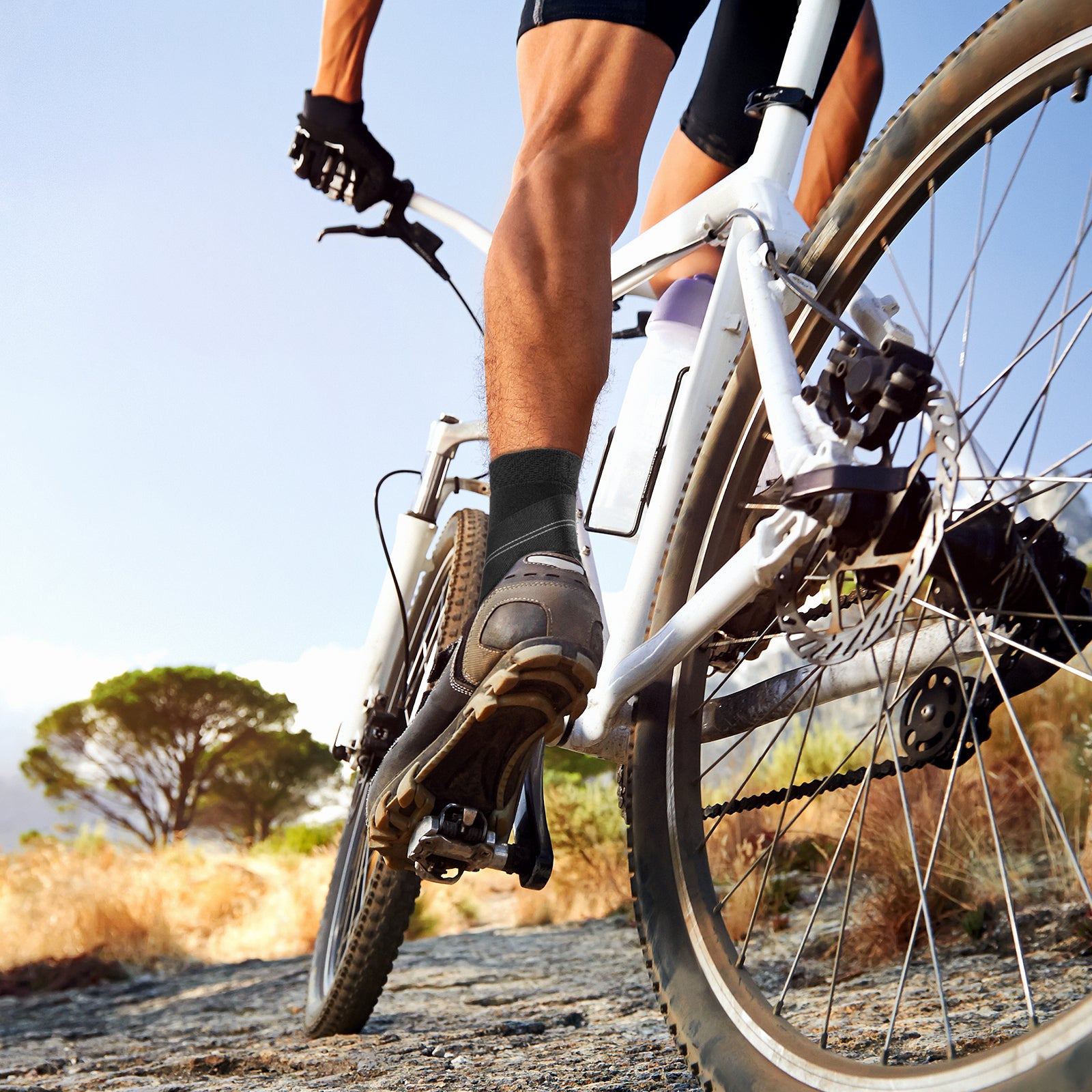 Outdoor cycling scene showing a person wearing a WHCOOL ankle compression sleeve with athletic shoes during a bike ride on a natural trail.
